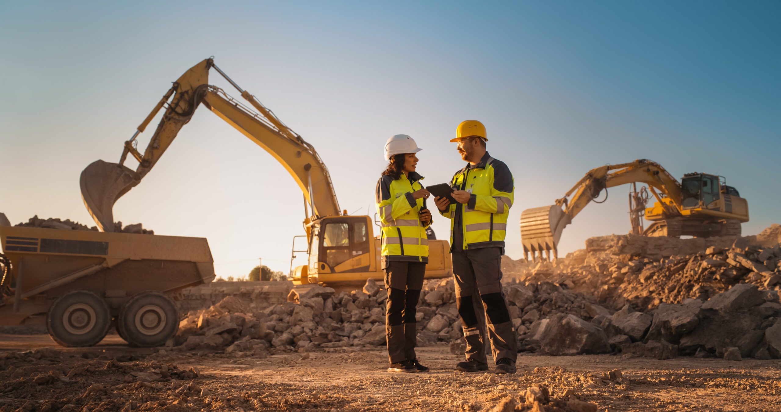 Hispanic Female Inspector Talking to Caucasian Male Land Development Manager With Tablet On Construction Site Of Real Estate Project. Excavators Preparing For Laying Foundation. Hot Sunny Day